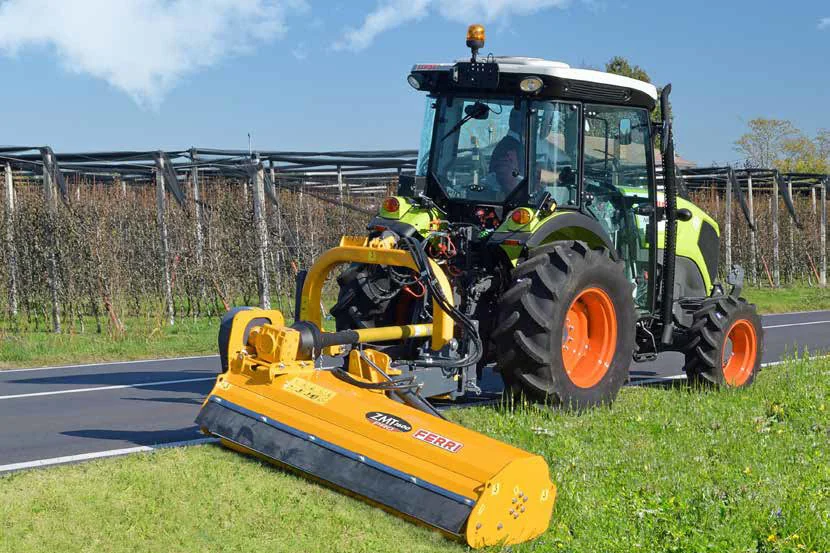 Tractor verd amb una desbrossadora Ferri groga desplaçada al costat, sobre l'herba de la vora d'una carretera, amb el cel blau