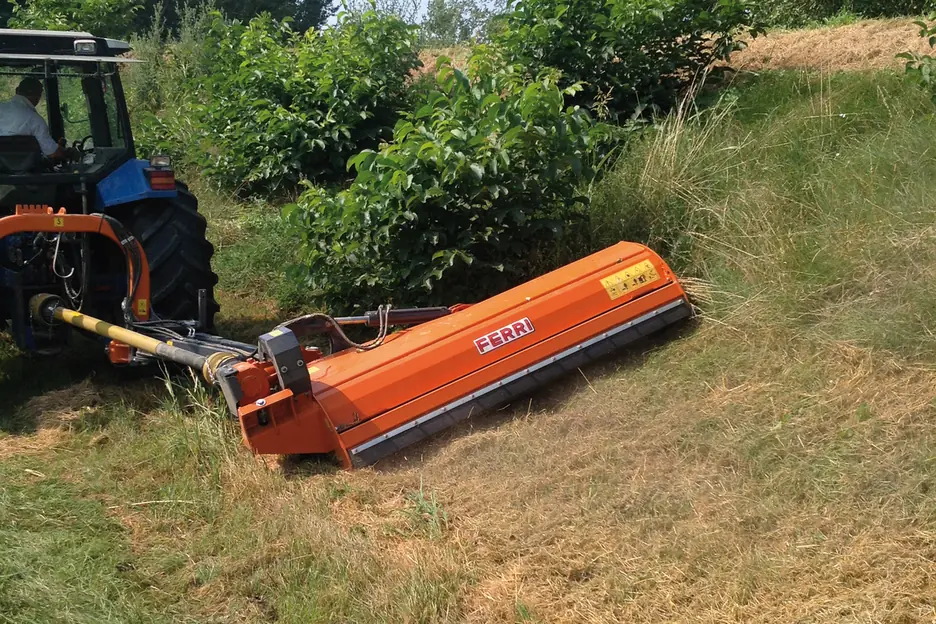 Desbrossadora lateral Ferri taronja segant un marge amb matolls, enganxada a un tractor blau