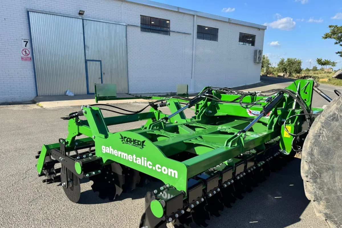 Grada ràpida Gaher amb l'adreça web del fabricant pintada al bastidor, davant d'una nau blanca i al costat de la roda d'un tractor