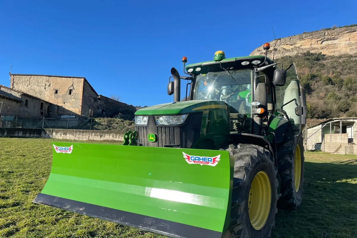 Pala polidozer Gaher verda muntada al davant d’un tractor verd, amb una masia i un cingle al fons