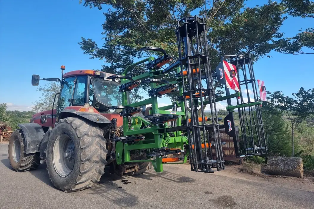 Xissel plegable Gaher amb les ales aixecades en vertical darrere d'un tractor vermell, sota un arbre