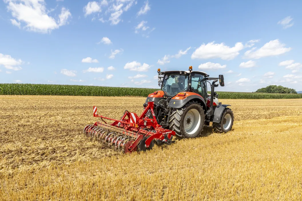 Grada de discos Horsch Joker CT suspesa als tres punts d'un tractor, treballant el rostoll amb un camp de blat de moro al fons