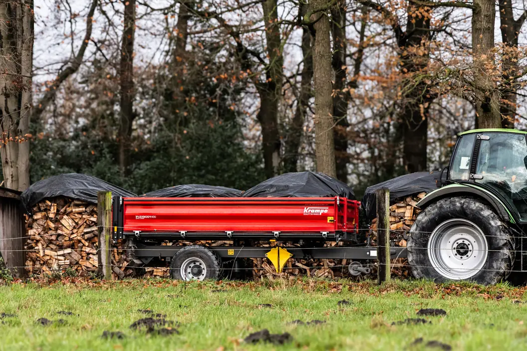 Remolc basculador Krampe FlexBody d'un eix aparcat al costat d'una pila de llenya tapada, amb la roda d'un tractor a la dreta