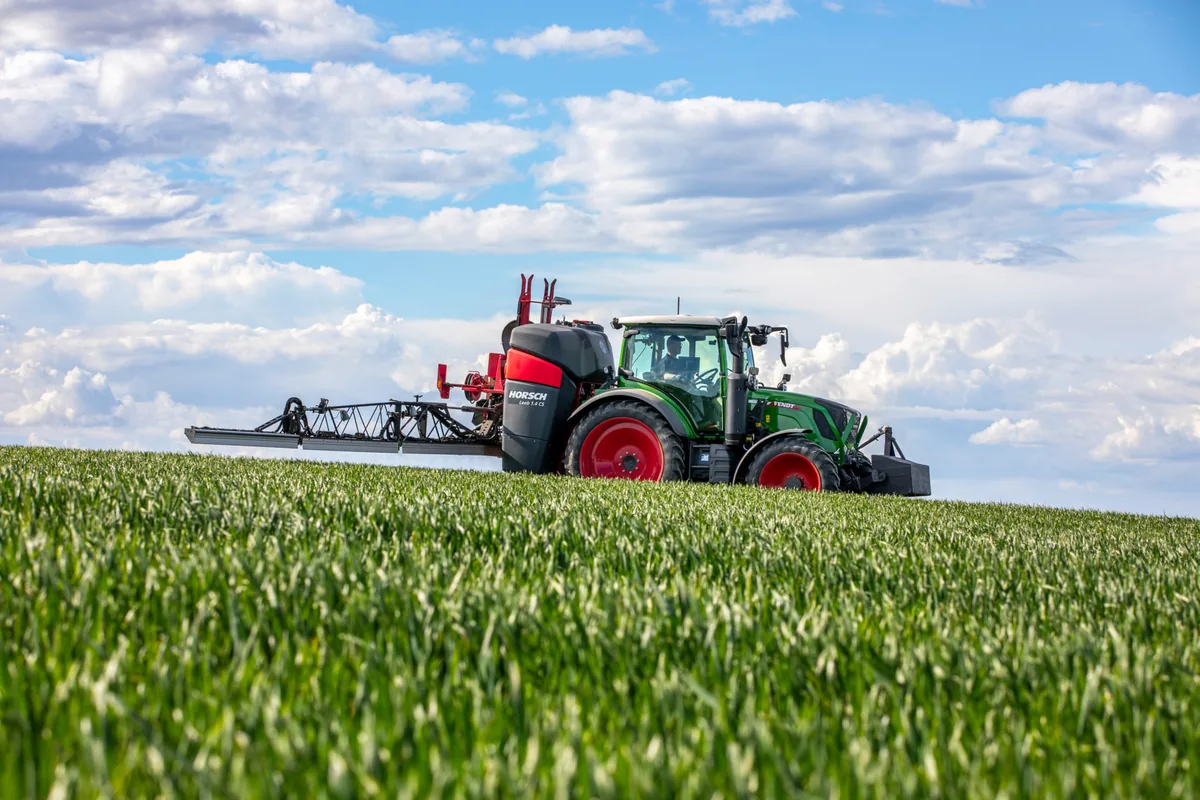 Polvoritzador Horsch Leeb CS suspès darrere d'un tractor verd, amb la barra desplegada sobre un camp de cereal