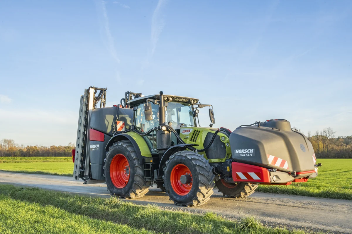 Tractor verd amb el dipòsit davanter Horsch Leeb 1.8 FT i un polvoritzador suspès amb la barra plegada al darrere, circulant per un camí de terra