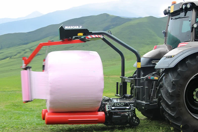 Encintadora Mascar 7100 muntada al darrere d'un tractor, amb una bala embolicada amb film rosa i turons verds al fons