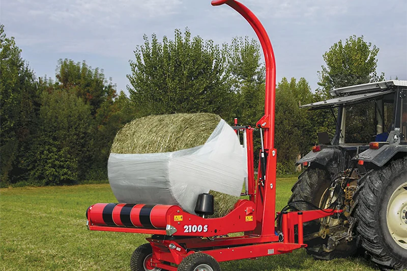 Encintadora Mascar 2100S remolcada amb una bala a la plataforma, enganxada a un tractor gris, amb arbres al fons