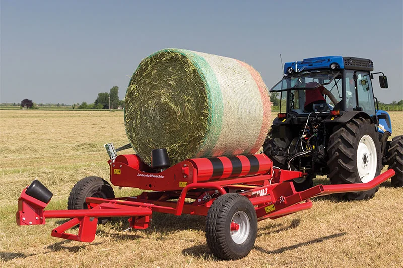 Encintadora Mascar 3100S remolcada amb una bala d'herba carregada, darrere d'un tractor blau, en un rostoll