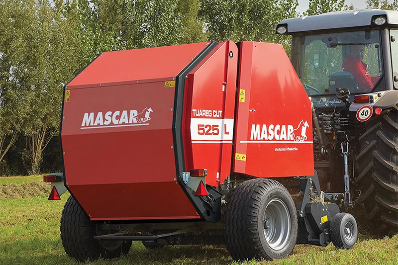 Rotoempacadora Mascar Tuareg Cut 525 de tres quarts pel darrere en un prat, enganxada a un tractor gris