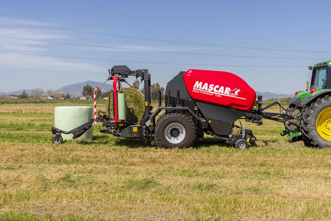 Rotoempacadora encintadora Mascar vermella i negra de perfil en un prat segat, enganxada a un tractor verd, amb una bala encintada deixada al costat