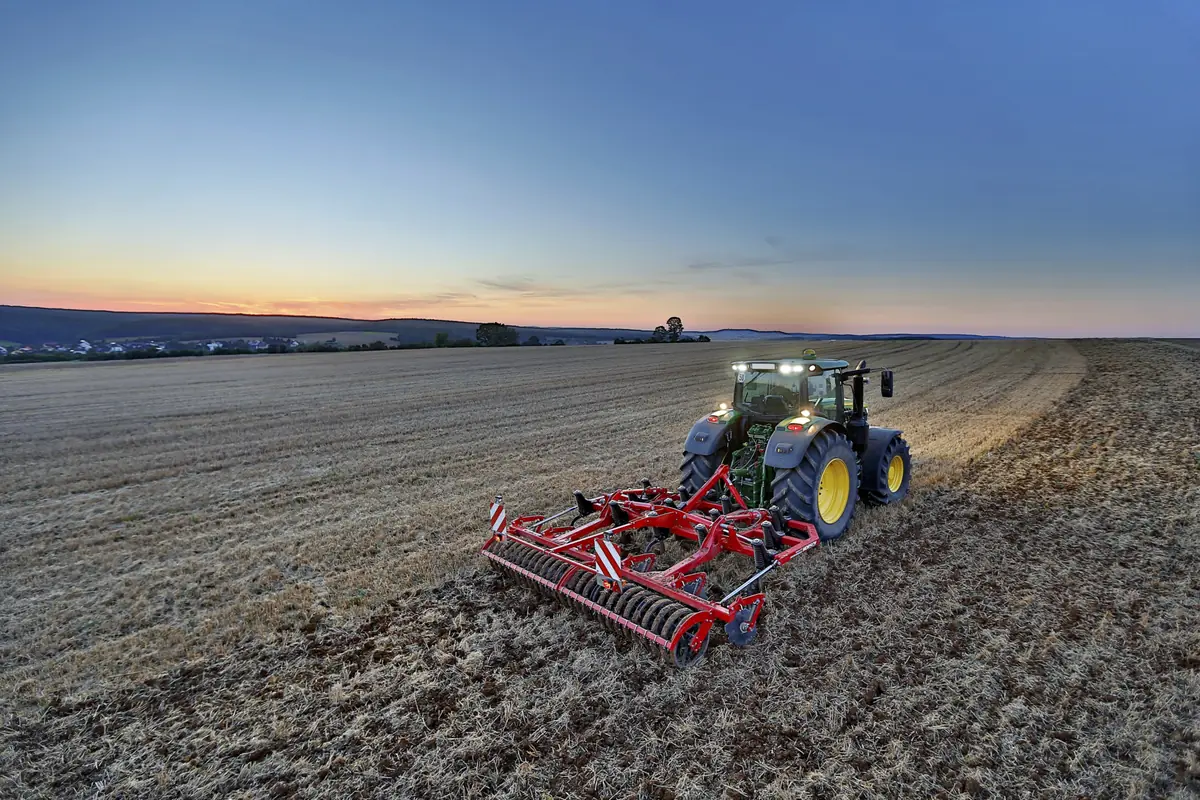 Vista aèria d'un cultivador Horsch Terrano FX suspès darrere d'un tractor amb els fars encesos, treballant el rostoll al capvespre