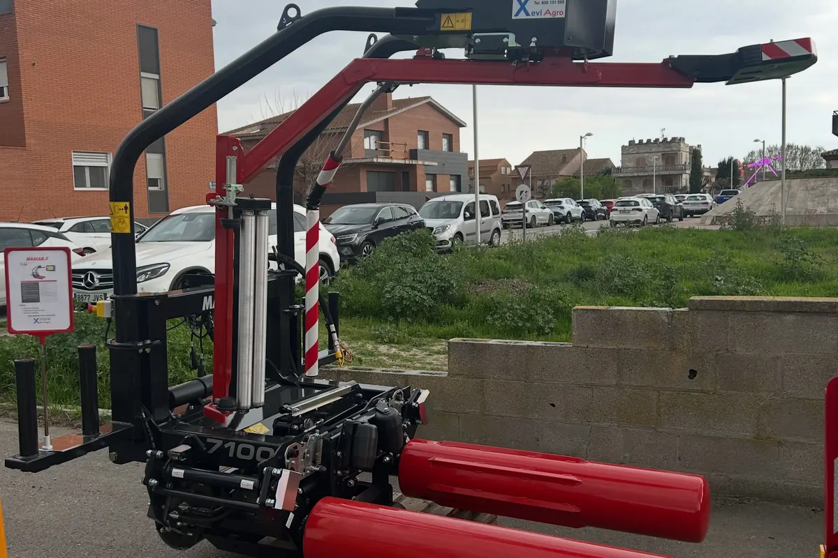 Encintadora de bales Mascar 7100 amb els dos braços vermells, aparcada al costat d'una tanca de blocs amb cases al fons