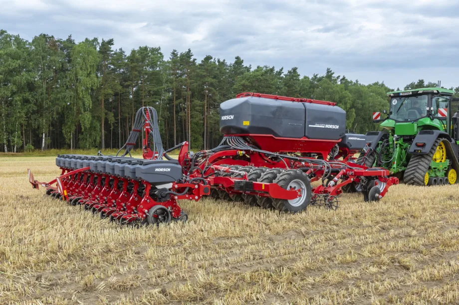 Sembradora de precisió Horsch Maestro sencera, amb la tremuja central i les unitats de sembra en filera, enganxada a un tractor d'erugues sobre un rostoll, amb un bosc de pins al fons