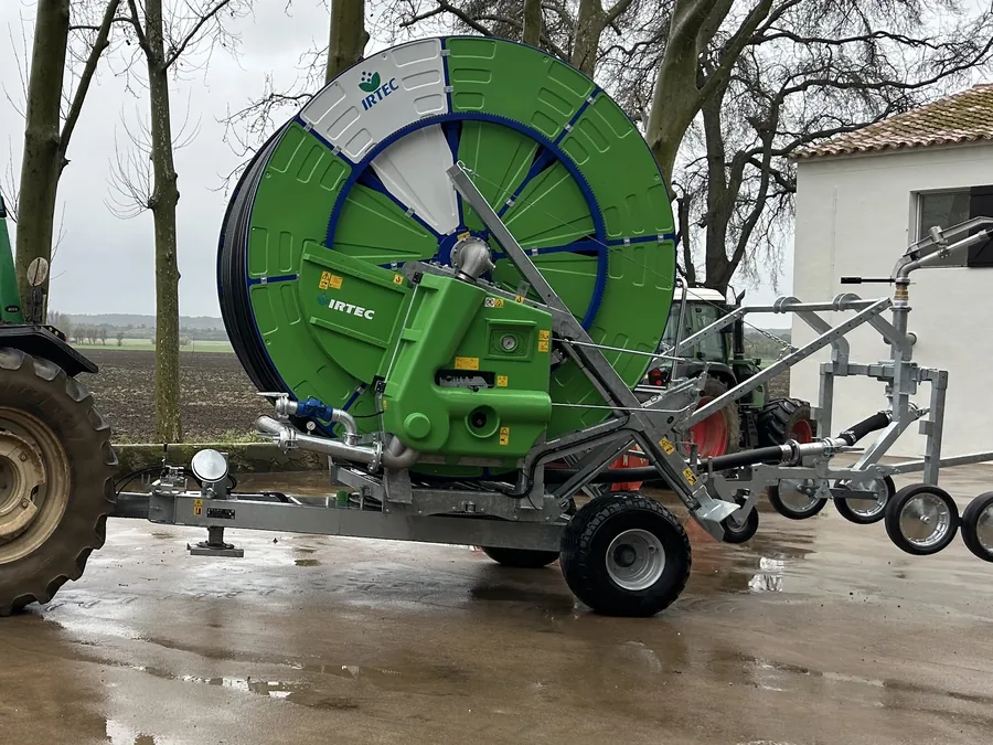Un enrotllador Irtec verd amb la mànega enrotllada al tambor, sobre paviment mullat, al costat d'un tractor i davant d'una casa