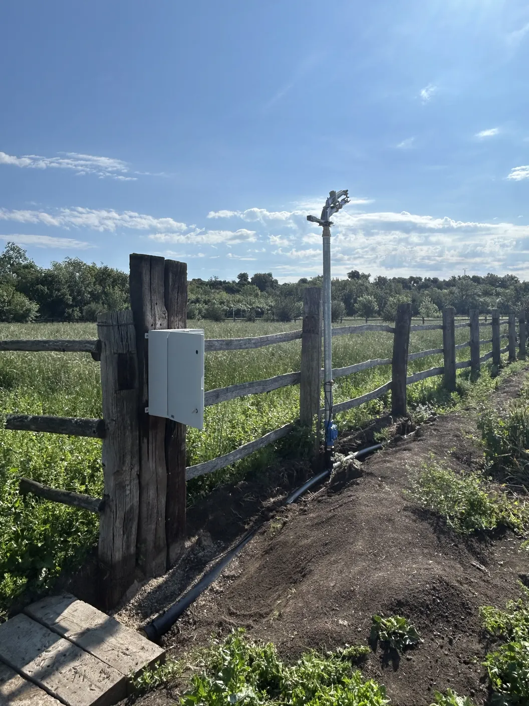 Un quadre de reg en un pal d'una tanca de fusta i, al costat, un aspersor damunt del seu tub amb l'electrovàlvula, vora un prat
